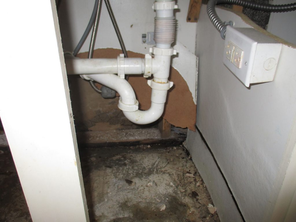 Sewage smell in house: under-sink cabinet with white PVC drain trap, electrical box and damaged, stained floor